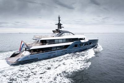 A large, sleek yacht with multiple decks and a tall mast is speeding across the choppy, gray waters of what appears to be an open sea or lake, with a cloudy, overcast sky in the background.