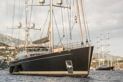 A sleek black yacht anchors offshore with a majestic three-masted schooner sailing in the background.