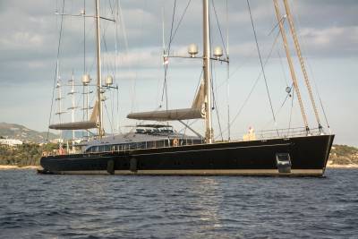 A sleek black sailing yacht with multiple masts glides through the calm, blue waters near a lush, green coastline.