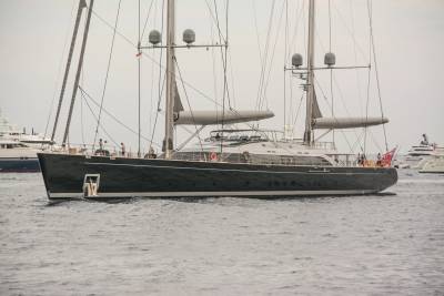 A sleek, dark yacht with tall masts glides through calm waters, accompanied by other vessels in the distance.
