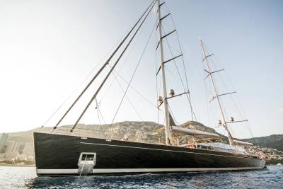 A majestic black sailing yacht glides through calm waters with a mountainous, coastal city in the background.