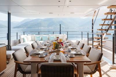 A beautifully set dining table awaits guests on a luxurious yacht deck, overlooking a serene bay with verdant mountains.