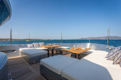 Relax on the spacious deck of a luxury yacht with comfortable seating and tables, surrounded by the sparkling blue sea and distant islands.