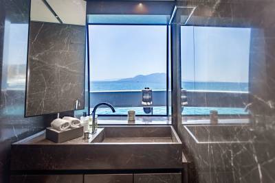 A modern bathroom with marble finishes offers a stunning view of the ocean and distant mountains through a large window.