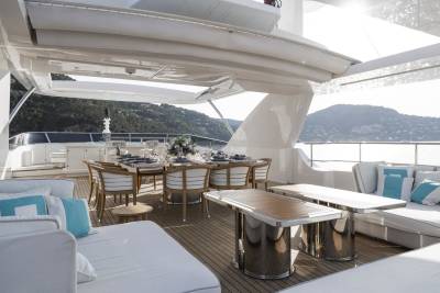 A beautifully set dining table awaits guests on the sun-drenched deck of a luxurious yacht overlooking a picturesque coastline.