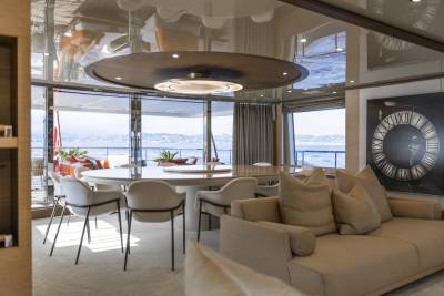 A luxurious yacht interior features a spacious dining area with a large round table and plush seating, overlooking a scenic ocean view.