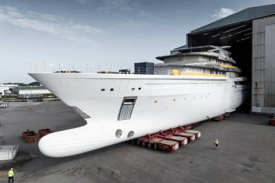 A colossal white yacht hull rests on a heavy-duty transporter outside a vast shipyard building.