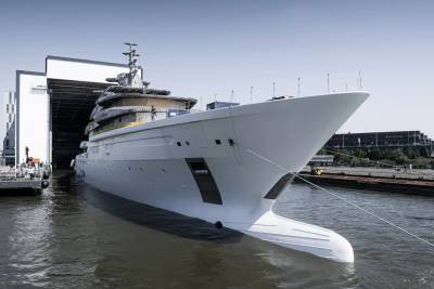 A massive white superyacht is being launched from a shipyard into the calm water under a clear blue sky.