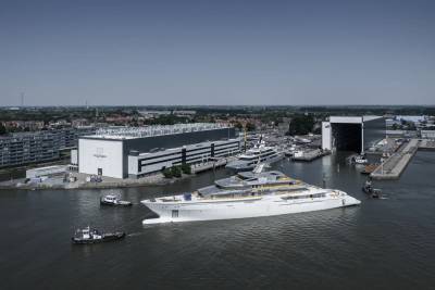 A massive white superyacht is being maneuvered by tugboats out of a shipyard under a clear blue sky.