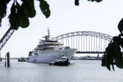 A massive superyacht navigates a waterway under a grand arched bridge, assisted by a small tugboat.