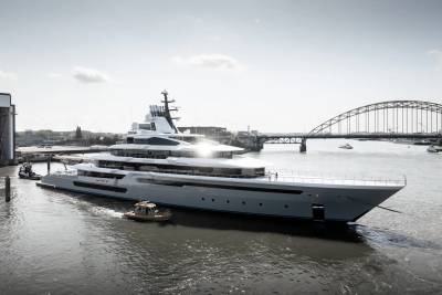 A colossal superyacht, the Leviathan, navigates a river under a bright sky, assisted by tugboats.