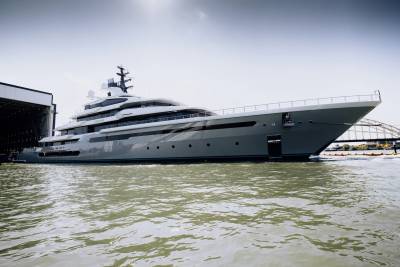 A massive, sleek grey yacht is docked in a shipyard, its modern design gleaming under a bright sky.