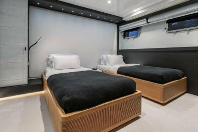 Two twin beds with dark gray blankets and white pillows are neatly made in a modern cabin with porthole windows.