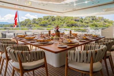 A beautifully set dining table on a yacht deck awaits guests with a stunning backdrop of lush green hills.