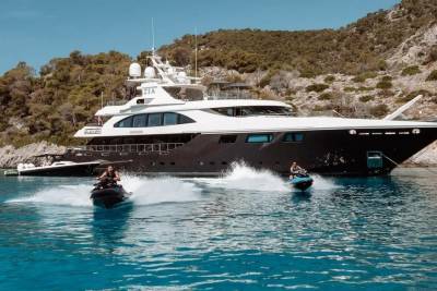 Two jet skiers race across the turquoise water near a luxurious yacht anchored by a rocky, tree-covered island.