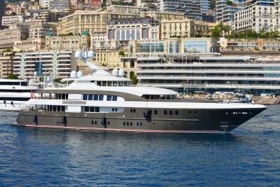 A sleek, dark-hulled superyacht glides through blue waters with a backdrop of Monaco's iconic cityscape.