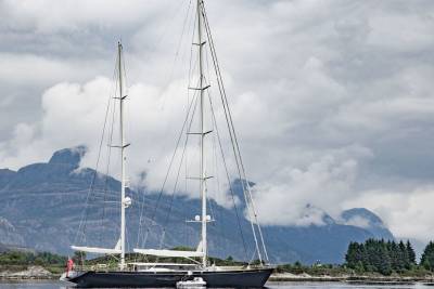 A majestic dark sailboat with towering masts glides through calm waters against a backdrop of misty, rugged mountains.