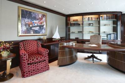 A luxurious yacht office features plush armchairs, a curved desk, and a wall of bookshelves filled with nautical-themed decor.