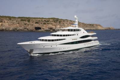 A luxurious white yacht glides through the deep blue sea near a rocky, vegetated coastline.