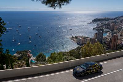 A black car with yellow accents drives along a scenic road overlooking the sparkling blue waters of Monaco, dotted with numerous yachts.