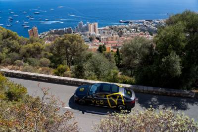 A sleek black car with yellow branding navigates a scenic road overlooking the azure waters and cityscape of Monaco.