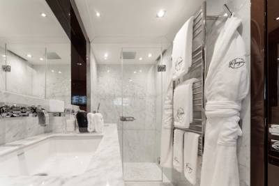 A spacious and well-lit bathroom with a marble-tiled floor, a vanity with a large mirror, and a towel rack holding white bathrobes.