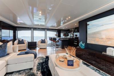 A luxurious and modern living room with large windows offering a stunning view of the ocean, featuring plush white sofas, a bar area, and a large television screen.