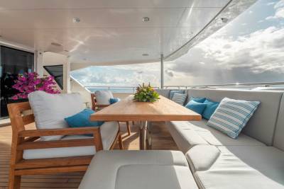A cozy outdoor seating area on a boat or yacht, with a wooden table, comfortable cushioned chairs, and vibrant floral arrangements, set against a backdrop of a cloudy sky and the open water.