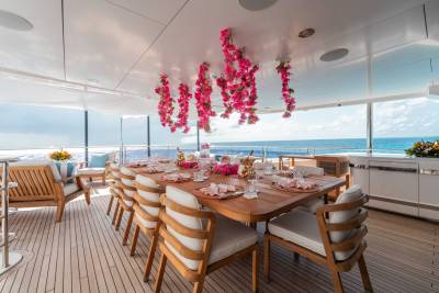 A beautifully decorated outdoor dining area on a yacht, with a long wooden table surrounded by comfortable chairs, adorned with vibrant pink floral arrangements and offering a stunning view of the ocean in the background.