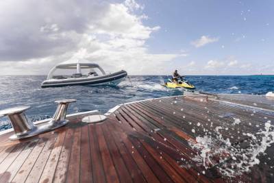 A wooden deck on a boat, with water splashing up, overlooks a person riding a jet ski in the distance against a cloudy sky.