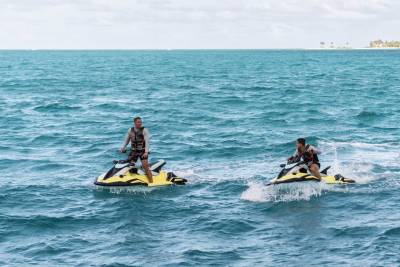 Two individuals are riding jet skis on the open ocean, with a coastal city visible in the background.