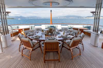 A beautifully set dining table awaits guests on the deck of a luxury yacht, overlooking a serene blue sea and distant mountains.
