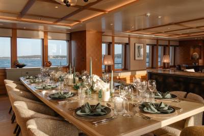 A beautifully set dining table awaits guests on a yacht, with a scenic ocean view through large windows.