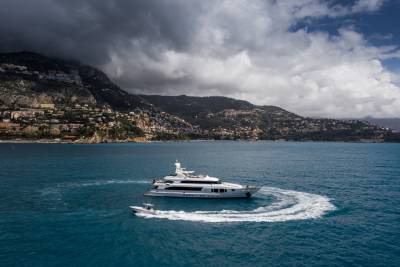 A luxurious white yacht cruises through the turquoise waters, leaving a swirling wake near a densely populated, verdant coastline under dramatic clouds.