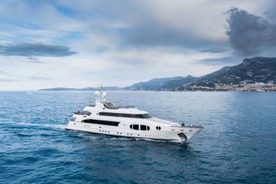 A luxurious white yacht cruises through the deep blue sea with a picturesque coastal town nestled in the mountains behind.
