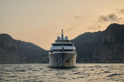 A majestic yacht glides through calm waters with rugged mountains rising behind it at sunset.