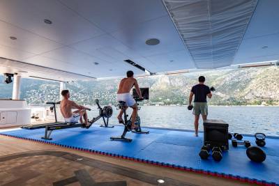Three men work out on a yacht's deck with rowing machines, stationary bikes, and free weights against a backdrop of a Mediterranean coastline.