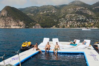 Friends relax on a floating deck with a pool and jet skis, overlooking a picturesque coastal town nestled in the mountains.