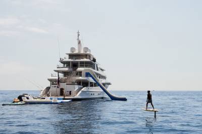 A person on a hydrofoil board glides across the calm blue sea near a large white yacht with a water slide.