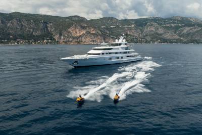 A luxurious white yacht cruises the azure waters, trailed by two jet skis creating white wakes against the dramatic mountainous coastline.