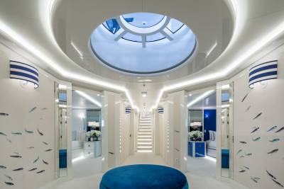 A luxurious yacht interior features a central blue ottoman, striped wall sconces, and playful fish wallpaper leading to a bright staircase.
