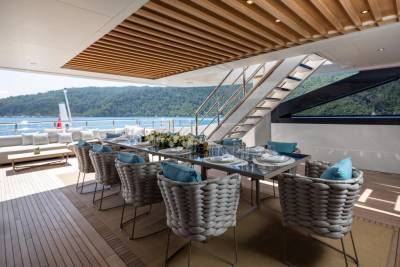 An elegant outdoor dining area on a yacht features a long table set for a meal with a backdrop of lush green hills and sparkling blue water.