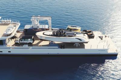 A sleek yacht support vessel carries a tender, a car, and jet skis across the shimmering blue ocean.