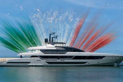 A sleek white yacht glides through the water as vibrant green, white, and red fireworks erupt behind it.