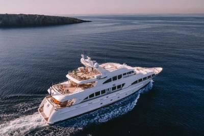 A luxurious white yacht cruises through the deep blue sea, leaving a foamy wake behind as a rocky island looms in the distance.