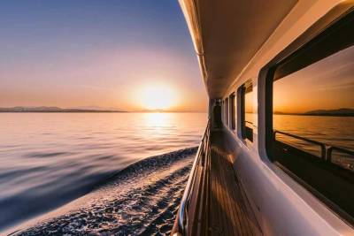 A yacht glides across calm waters at sunset, reflecting the warm hues of the sky and distant mountains.