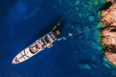 A luxurious yacht anchors near a rocky coastline, surrounded by crystal-clear turquoise waters and various water toys.