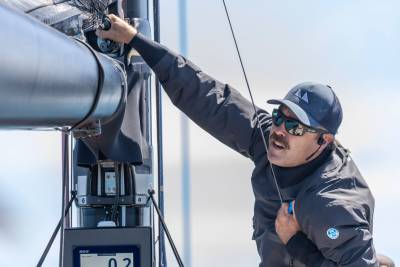 A sailor adjusts rigging on a boat, his face obscured by sunglasses and a cap.