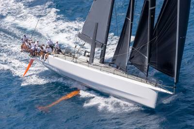 A sleek white sailboat with dark sails cuts through the blue ocean, its crew visible on deck.