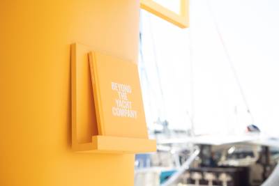 A bright yellow book titled "Beyond the Yacht Company" is displayed on a matching shelf against a blurred backdrop of yachts.